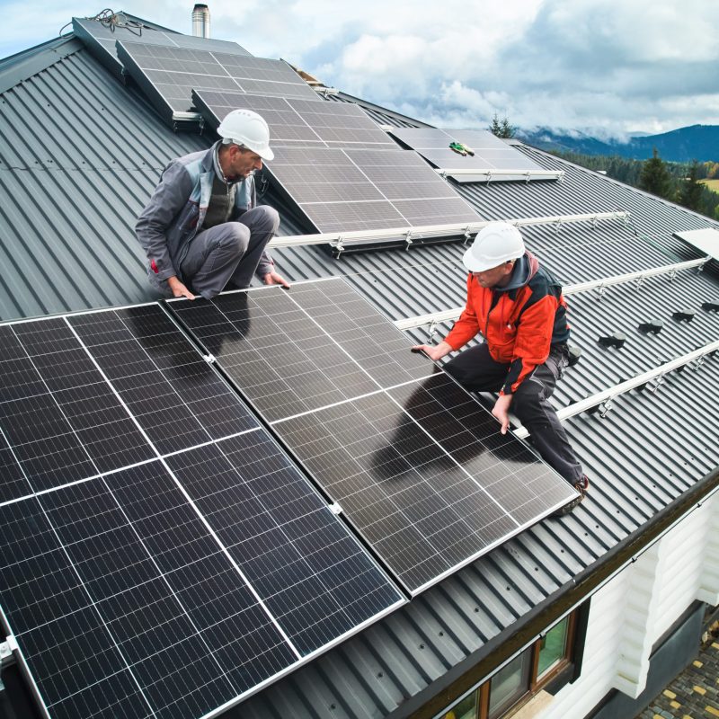 Workers building photovoltaic solar module station on roof of house. Men electricians in helmets installing solar panel system outdoors. Concept of alternative and renewable energy. Aerial view.